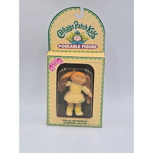 May include: A Cabbage Patch Kids poseable figure in its original packaging. The doll has orange hair, a white dress, and yellow shoes, holding a small brown teddy bear. The box features the text First Edition and Hello, My Name Is Jennifer Lillian.