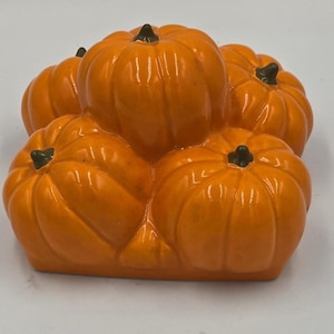 May include: A decorative ceramic sculpture featuring a cluster of five glossy orange pumpkins arranged together. Each pumpkin has a smooth, rounded surface with vertical ridges and a small, dark green stem at the top center of each individual gourd.