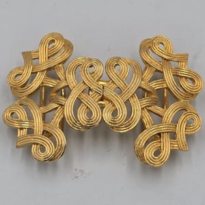 May include: A gold-toned metal belt buckle featuring a symmetrical design of interconnected, swirling loops and linear ridges. The rectangular-shaped accessory displays a continuous, woven pattern across its entire surface with a polished finish.