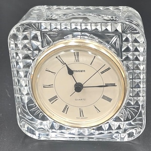 May include: A square crystal desk clock with a faceted, diamond-cut pattern. The clock face is cream-colored with black Roman numerals, black hands, and the text "STAIGER," "QUARTZ," and "WEST GERMANY" printed in black.