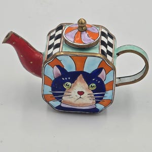 May include: A small, decorative metal teapot featuring a hand-painted cat face on the side. The teapot has a red spout, a black and white checkered pattern on the sides, and a lid with orange and purple stripes. It has a brass-toned metal trim.