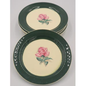 May include: A stack of round ceramic plates featuring a wide, dark forest green rim and a cream-colored center. Each plate is decorated with a single pink rose illustration with green leaves centered on the cream background.