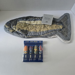 May include: A fish-shaped quilted kitchen oven mitt with a tan and grey spotted pattern, accompanied by a set of four corn picks in a blue box. The mitt tag reads kitchen OVEN MITT 5.75 x 16.25 inches. The corn picks feature colorful fish designs.