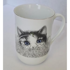 May include: A white ceramic mug featuring a detailed black and white illustration of a kitten with bright blue eyes peeking through tall grass. The kitten has a small green caterpillar on a blade of grass above its head. The mug has a handle.