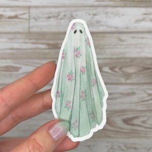 Könnte beinhalten: Ein gestanzter Aufkleber mit einem Geisterdesign. Der Geist ist in ein hellgrünes Laken gehüllt, das mit rosa Blumenmustern verziert ist. Der Aufkleber hat einen weißen Rand und ein mattes Finish.