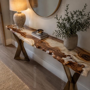 May include: A long console table featuring a natural live-edge wood slab surface with integrated translucent white resin sections. The table is supported by two metallic gold-toned X-shaped legs, creating a distinct contrast between the organic wood grain and the modern base structure.