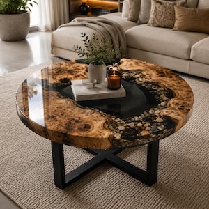 Peut inclure: Une table basse ronde composée d'un plateau en bois brut à bord naturel et d'un centre en résine brillante incrusté de galets. La table repose sur un piètement en croix en métal noir. Un livre blanc, une plante et une bougie sont posés dessus.