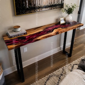May include: A long console table featuring a natural wood slab top with a central river of deep magenta resin. The table is supported by two black metal U-shaped legs. The surface holds a stack of books, a small white container, and a vase.