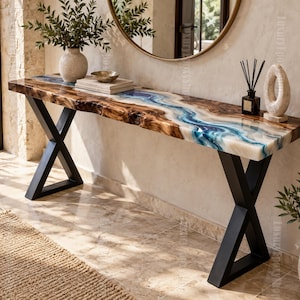 Può includere: Un lungo tavolo console caratterizzato da un piano in legno naturale con bordo irregolare e un inserto centrale in resina blu, bianca e traslucida. Il tavolo è sostenuto da due gambe in metallo nero a forma di X e ha una finitura lucida.