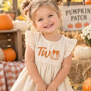 May include: A cream-colored sleeveless dress with ruffled shoulders, featuring orange embroidery on the chest that reads "Two" next to a pumpkin graphic with a bow. The dress has a gathered waist and a simple round neckline.