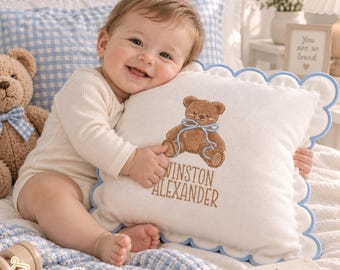 May include: A square white throw pillow with a scalloped light blue edge, featuring an embroidered brown teddy bear with a light blue ribbon and the name Winston Alexander in brown text, held by a baby in a cream-colored long-sleeve bodysuit.