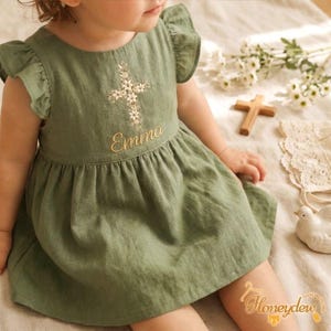 May include: An olive green dress with ruffled sleeves, featuring an embroidered floral cross design on the chest above the name Emma stitched in gold thread. The dress has a round neckline and a gathered skirt that falls from the bodice.
