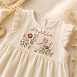 May include: A cream-colored sleeveless dress featuring ruffled shoulders and a round neckline. The chest area is decorated with embroidered flowers, green stems, and a white goose, with the name Amelia stitched in pink cursive script above.