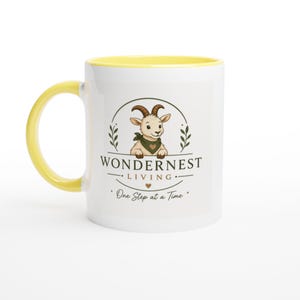 Könnte beinhalten: Ein weißer Keramikbecher mit einem leuchtend gelben Henkel und Innenrand. Auf der Vorderseite befindet sich eine Grafik einer Cartoon-Ziege mit grünem Halstuch, umrahmt von dem Text WONDERNEST LIVING und One Step at a Time.