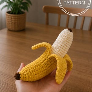 Puede incluir: Un juguete de plátano tejido a ganchillo con una cáscara amarilla brillante, un centro de fruta color crema y puntas de color marrón oscuro. El artículo se presenta como un patrón PDF, con el texto PDF PATTERN visible en un gráfico circular.