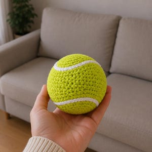 Puede incluir: Una pelota de ganchillo de color verde lima, de forma redonda, con una franja de hilo blanco que se curva alrededor de la superficie, imitando el aspecto de una pelota de tenis. La textura se crea mediante puntos de hilo visibles y uniformes.