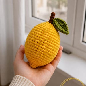 May include: A yellow crocheted lemon toy with a brown stem and a single green leaf. The lemon has a textured, knotted surface created by the crochet stitches. The item is labeled with the text PDF PATTERN in a gold circular graphic.
