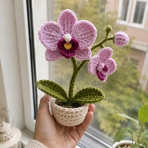 Może przedstawiać: Szydełkowana orchidea z dwoma dużymi jasnoróżowymi kwiatami z ciemnomagentowymi i żółtymi środkami, małym różowym pąkiem i dwoma zielonymi liśćmi. Roślina znajduje się w małej, okrągłej, kremowej szydełkowanej doniczce.