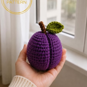 Puede incluir: Una ciruela de ganchillo morada con un tallo marrón y una hoja verde. El texto PDF PATTERN es visible en la esquina superior izquierda. La fruta tiene una forma esférica texturizada con una hendidura vertical central, hecha de hilo.