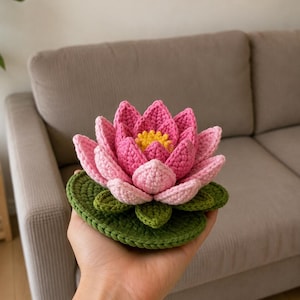 Peut inclure: Une fleur de lotus crochetée à la main avec des pétales roses superposés, un centre jaune vif et une base circulaire en forme de nénuphar vert foncé. La fleur est réalisée avec des points de laine visibles, créant un design texturé en relief.