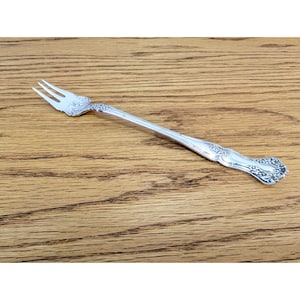 May include: A silver-toned metal cocktail fork featuring three tines and an elongated handle. The handle and the base of the tines are decorated with an embossed floral and leaf pattern that extends along the length of the slender stem.