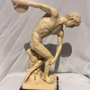 Op de afbeelding: Een crèmekleurig beeldhouwwerk van een discuswerper in een dynamische atletische houding, vastgelegd in beweging met een schijf in de rechterhand. Het figuur rust op een boomstomp en staat op een rechthoekige voet van zwart marmer.