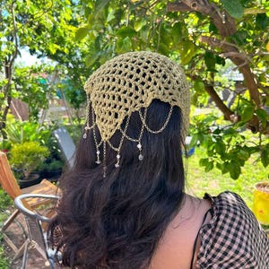 May include: A sage green crochet head covering featuring a triangular shape with an open-knit pattern. The edges are adorned with delicate gold-toned metal chains that drape down, accented with small clear beads and tiny white pearls.