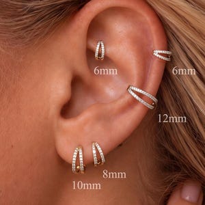 May include: A set of gold-toned hoop earrings featuring clear stones, displayed on an ear. The jewellery includes various sizes labelled as 6mm, 8mm, 10mm, and 12mm, showcasing double-band designs with embedded crystals along the outer edges.