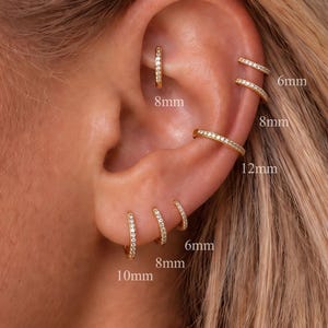 May include: A set of gold hoop earrings with embedded clear stones, worn in multiple ear piercings. The hoops are labelled with sizes 6mm, 8mm, 10mm, and 12mm, indicating their respective diameters for various placements along the ear cartilage and lobe.