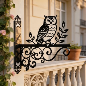 Puede incluir: Un soporte de pared de metal negro que presenta una silueta decorativa de un búho posado en una rama. El soporte incluye intrincados diseños de volutas debajo de la barra horizontal y una placa de montaje vertical con una flor de lis.