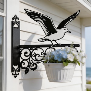 Puede incluir: Un soporte de pared de metal negro para plantas con la silueta de una gaviota con las alas extendidas. El soporte tiene un diseño de volutas ornamentadas y sostiene una maceta colgante blanca, redonda y acanalada con flores azul claro y blancas.