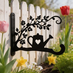 May include: A black metal wall-mounted plant hanger bracket featuring a decorative silhouette of a watering can with a heart-shaped cutout, accented by a branch design with leaves and small flowers, extending from a vertical mounting plate.