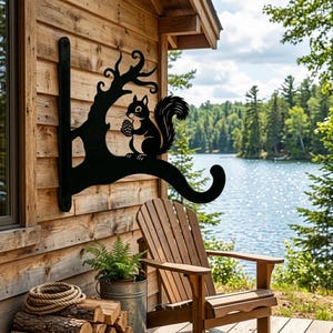 May include: A black metal wall-mounted bracket featuring a silhouette of a squirrel perched on a tree branch holding a pinecone. The bracket has a curved hook at the end for hanging items and is attached to a vertical mounting plate with screw holes.