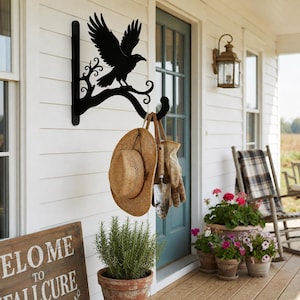 May include: A black metal wall-mounted hook featuring a silhouette of a bird perched on a branch. The hook supports a tan straw hat and a pair of tan work gloves, with a small metal hand trowel hanging from the center of the hook structure.