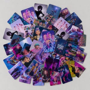 Op de afbeelding: Een verzameling rechthoekige ruilkaarten met kleurrijke illustraties van personages uit K-Pop Demon Hunters. De kaarten tonen diverse scènes met levendige paarse, blauwe en roze verlichting, met uiteenlopende geanimeerde figuren.