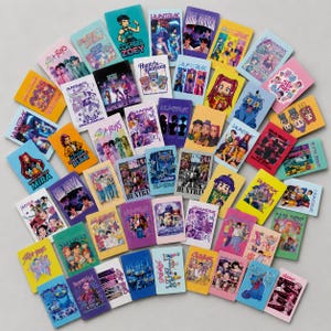 May include: A collection of rectangular trading cards featuring colorful anime-style illustrations of characters, groups, and creatures. The cards display various titles including Huntrix, Saja Boys, and Kpop Demon Hunters, with vibrant backgrounds.
