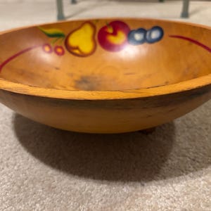 May include: A round wooden bowl with a light brown finish and a smooth interior. The inner rim features a hand-painted design of a yellow pear, a red apple, and two blue berries, all connected by a thin red line that circles the bowl's interior.