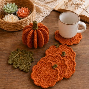 May include: A set of crocheted items including a stuffed orange pumpkin, four pumpkin-shaped coasters, and one green leaf-shaped coaster. A white ceramic mug sits on one coaster, while a woven basket holds three succulent-shaped candles.