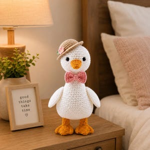 May include: A white crocheted duck toy featuring a tan hat with a small pink flower, a pink bow tie, and orange feet. The duck has black bead eyes and an orange beak. The toy is standing upright with a rounded body and two small wings.