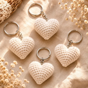 May include: Four off-white heart-shaped keyrings made of crocheted yarn. Each heart is attached to a silver-toned metal split ring by a short metal chain. The texture of the crochet stitches is visible on the surface of each heart.