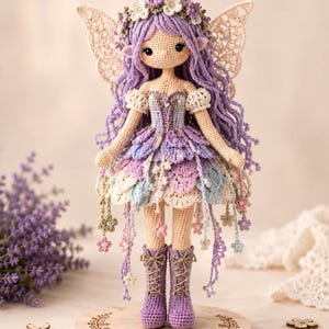 May include: A crocheted doll with long lavender hair, a floral crown, and lace wings. The doll wears a layered petal skirt in pastel shades of purple, blue, and cream, with dangling flower accents and purple boots. It stands on a wooden base.