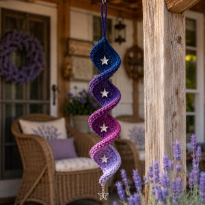 May include: A spiral-shaped crochet hanging ornament featuring a colour gradient from dark blue to light purple. The piece includes five silver-toned star charms attached at intervals along the centre, with a loop at the top for hanging.