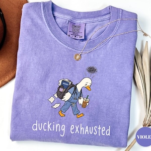 May include: A violet short-sleeve t-shirt featuring a graphic of a duck wearing blue scrubs, a stethoscope, and a backpack filled with papers and a book. The duck holds an iced coffee. The text "ducking exhausted" is printed below the graphic.