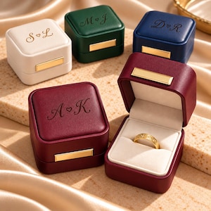 May include: Four square ring boxes in white, dark green, navy blue, and burgundy, each featuring a gold-tone metal clasp and engraved initials with a heart. One burgundy box is open, revealing a gold ring inside on a cream velvet insert.
