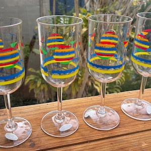 May include: A set of four clear glass goblets featuring a colourful graphic print of horizontal striped circles in red, yellow, blue, and green. Each glass has a slender stem and a circular base with a small white sticker attached to the bottom.
