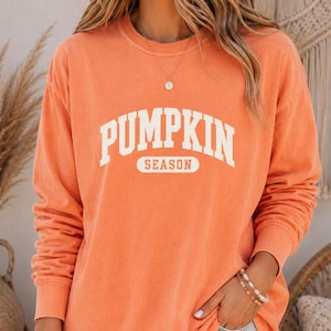 Op de afbeelding: Een sweatshirt met lange mouwen en ronde hals in een vervaagde oranje kleur met de witte tekst PUMPKIN SEASON in een collegiaal lettertype op de borst. Het kledingstuk heeft een relaxte pasvorm met geribbelde boorden aan de mouwen.