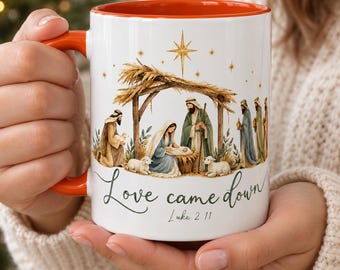 May include: A white ceramic mug with an orange interior and handle, featuring a colorful nativity scene illustration, a gold star, and the text "Love came down" followed by "Luke 2:11" in a dark green script font on the front surface.