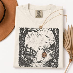 May include: A cream-colored t-shirt featuring a black and white ink-style illustration of a ghost in a sheet carrying a pumpkin. The scene depicts a path through trees, a wooden fence, and flying bats. The shirt has a Comfort Colors brand label.