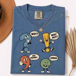 Può includere: Una t-shirt blu con una stampa grafica di quattro segni di punteggiatura antropomorfi. Un punto interrogativo blu dice WHY?, un punto esclamativo giallo dice WHY NOT!, una virgola rossa dice ONE MOMENT... e un punto verde dice ENOUGH.