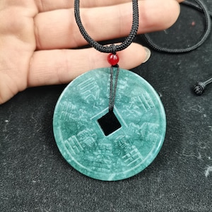 May include: A circular green stone pendant featuring carved Chinese characters and trigram symbols. The pendant has a square centre hole and is suspended from a black braided cord with a single small red bead accent above the stone piece.